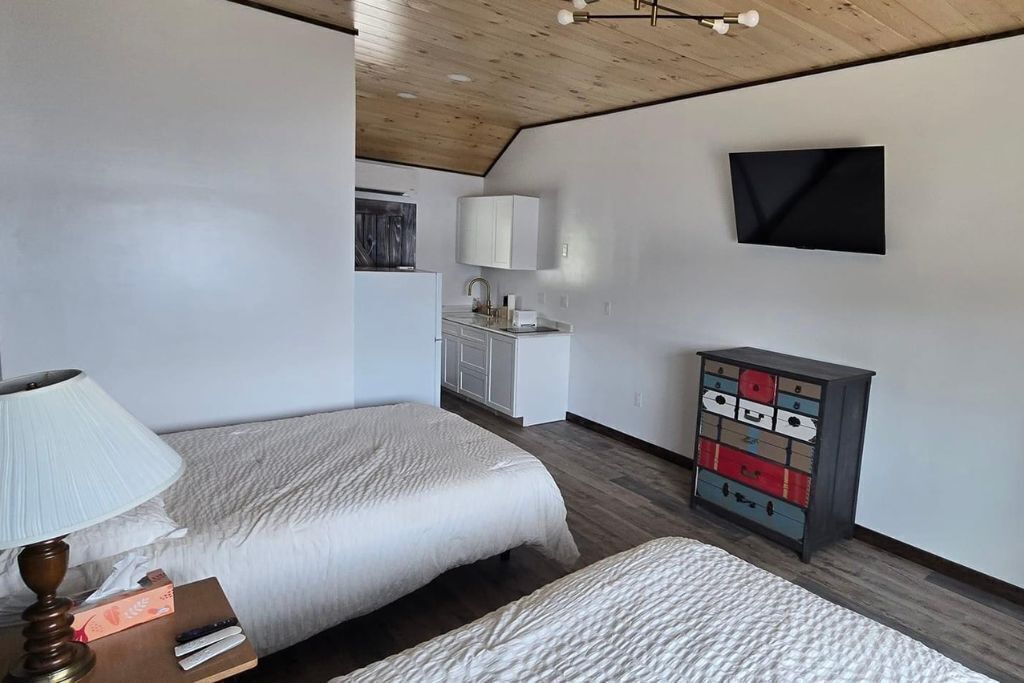 Une chambre avec deux lits, une table de chevet avec une lampe, une commode avec des tiroirs, un téléviseur fixé au mur et, au fond, une petite cuisine.