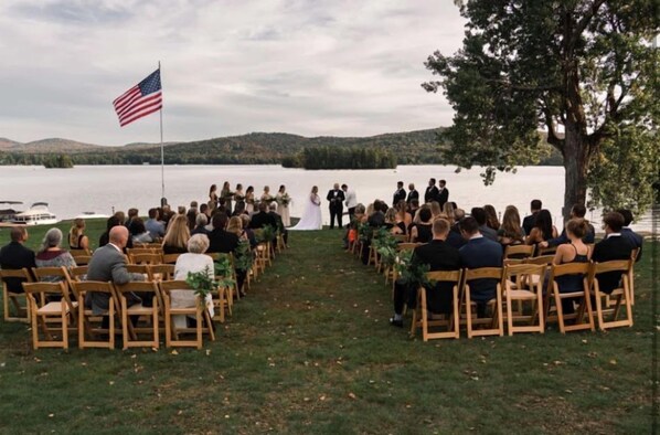 A wedding ceremony outdoors with guests seated on wooden chairs facing a couple at the front.