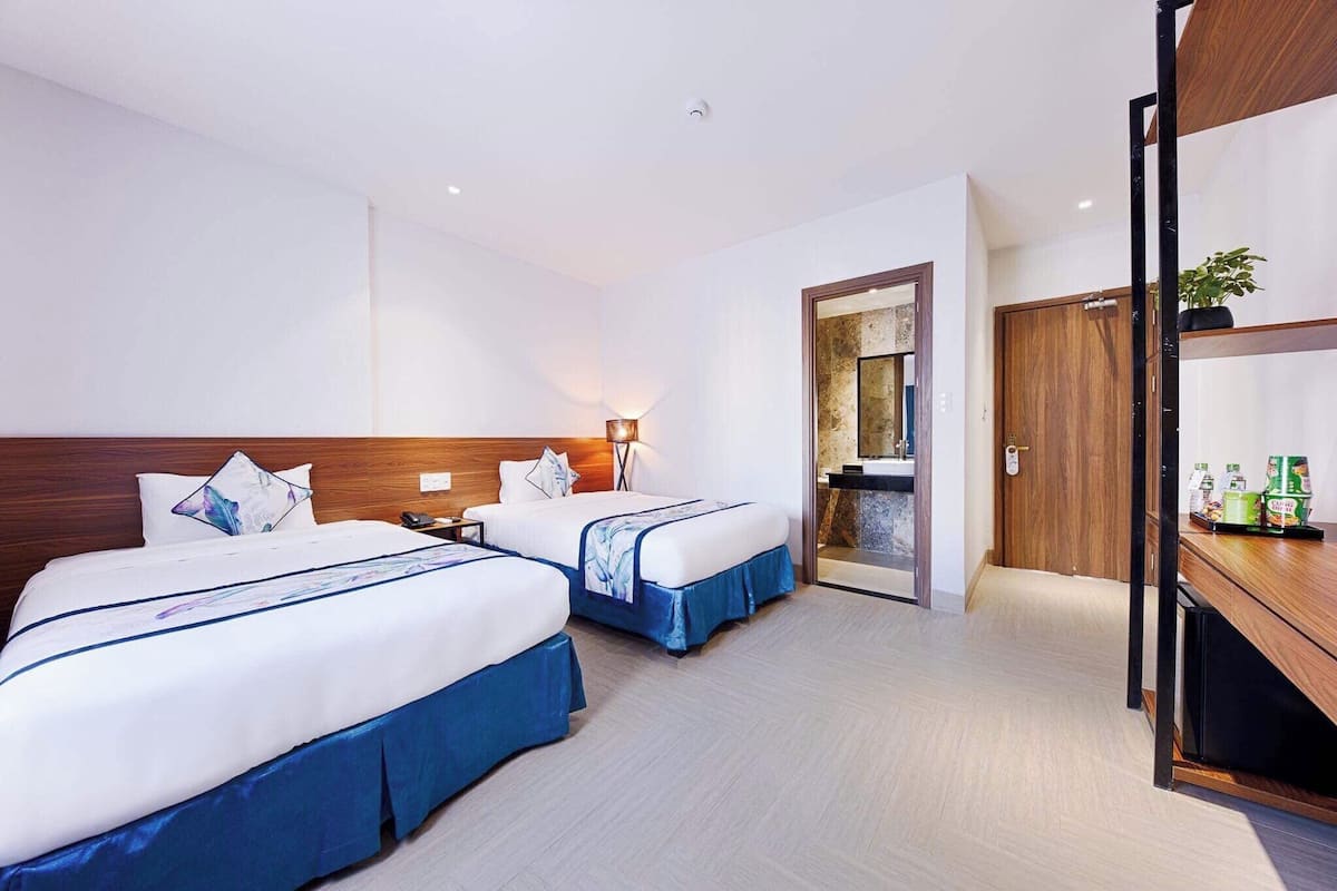 A hotel room with two beds, a wooden headboard, a nightstand, a lamp, a mirror, a door, and a small shelf with bottles.