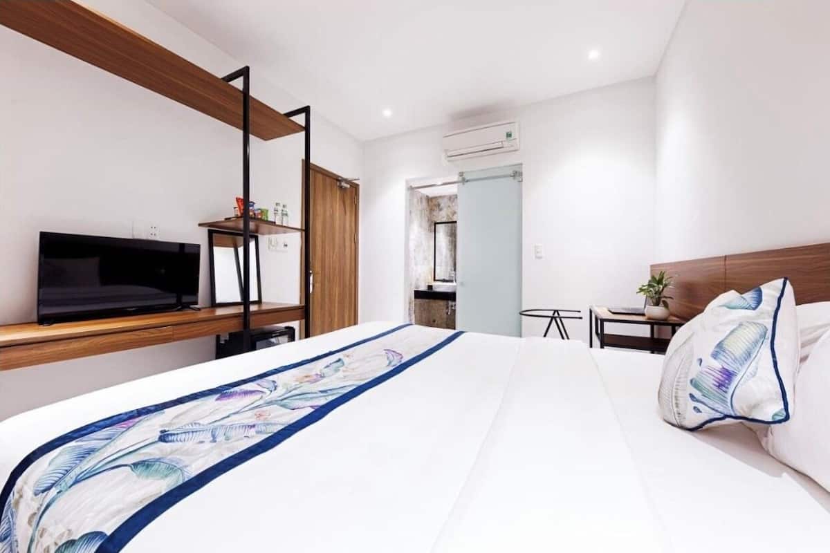 A modern hotel room with a large bed, a TV on a wooden shelf, a small table with a plant, and a bathroom visible through an open door.