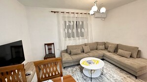 A living room with a sectional sofa, a TV, a wooden chair, a small round table, and a window with sheer curtains.