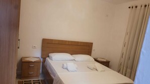 A neatly made bed with white linens and pillows, a wooden headboard, a bedside table with a telephone and a small decorative item, and a wooden nightstand with drawers.