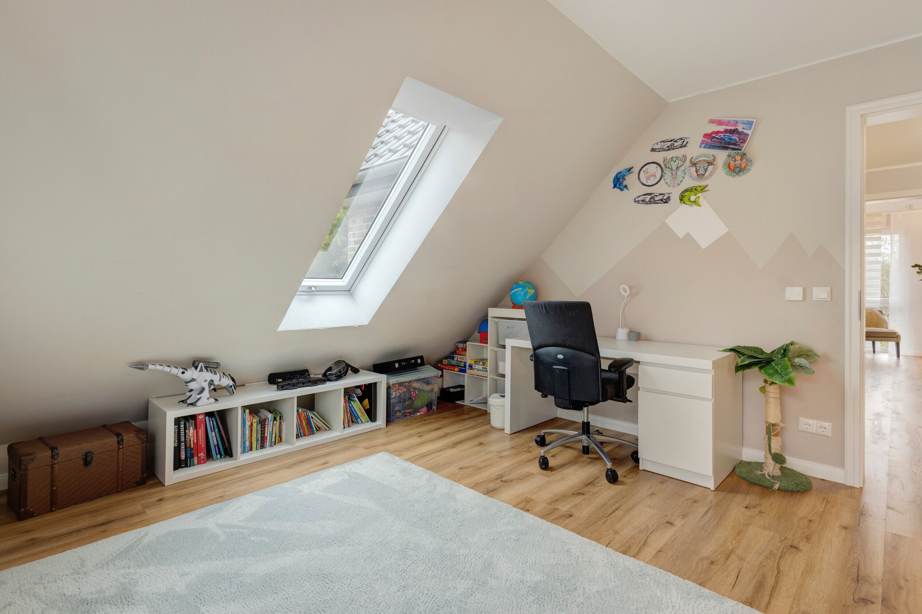 Una habitación con techo inclinado, escritorio, silla, estantería, un globo terráqueo y una planta.