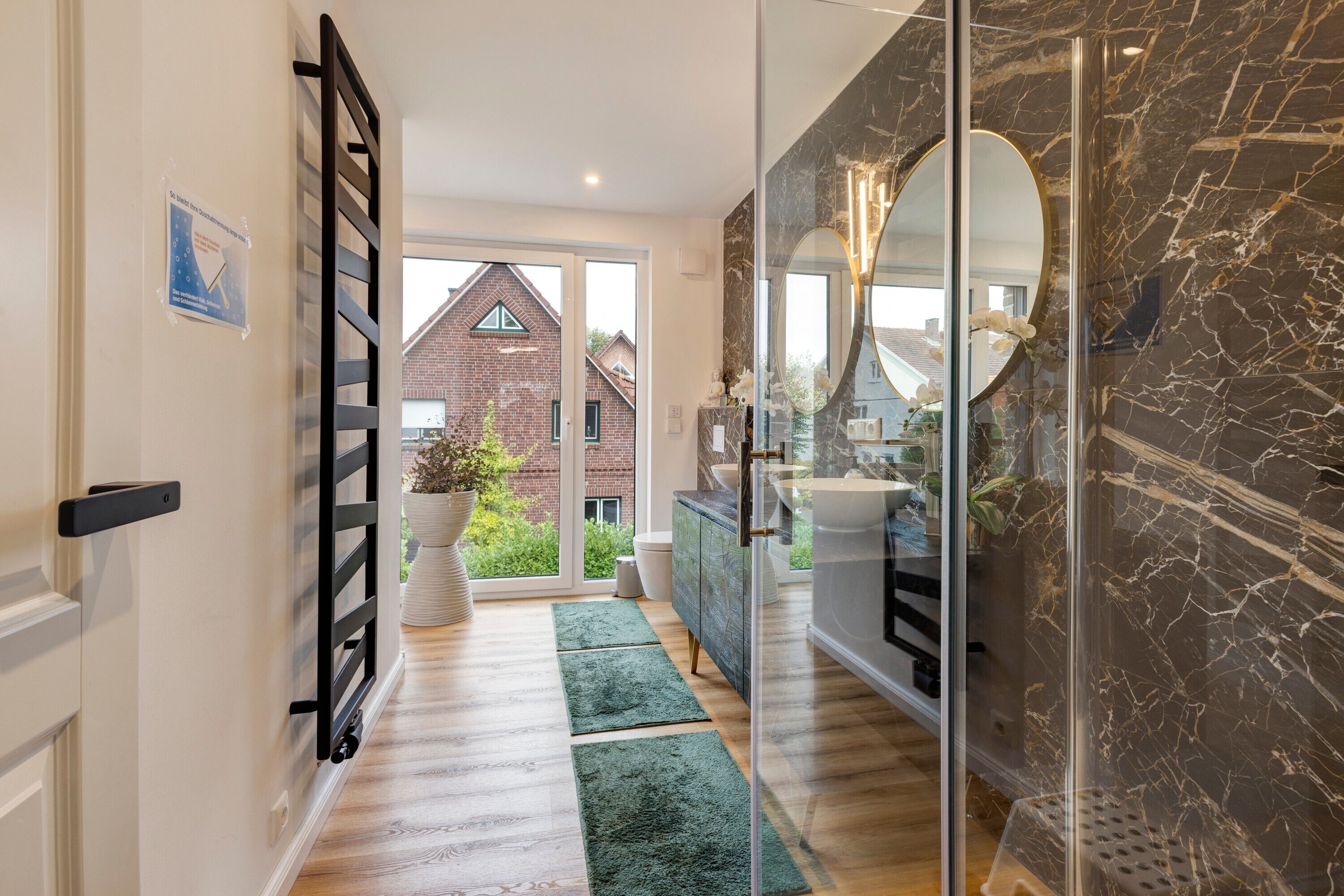 Ein modernes Badezimmer mit einer Glasdusche, einer Marmorwand und Blick auf ein Backsteinhäuschen durch die Tür.