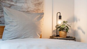 Una cama bien hecha con un almohadón blanco, una mesita de noche con una cesta tejida que contiene una planta y una lámpara moderna.