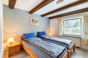 Un dormitorio con dos camas, cabeceros de madera, una mesita de noche con lámpara, un cuadro enmarcado en la pared y una ventana con cortinas.