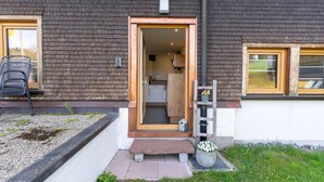 A residential building with a dark brick exterior, a wooden door leading to a kitchen, and a small garden area with a metal chair and a concrete planter.