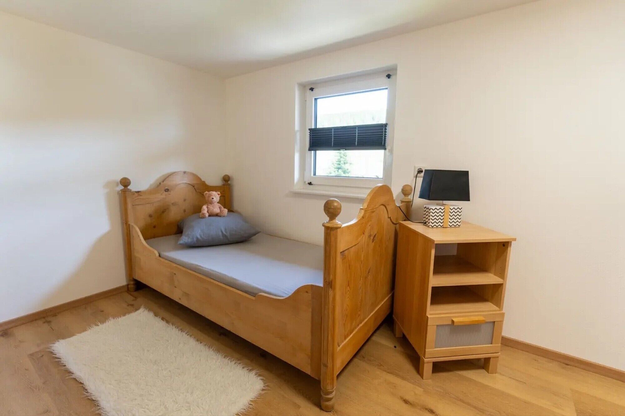Une petite chambre avec un lit simple, une tête de lit en bois, une table de chevet avec une lampe et une fenêtre équipée de stores.