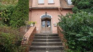 A two-story house with a brick staircase leading to a wooden door, surrounded by lush greenery. - Apartment 'Wohnen Im Alten Rittergut' with Private Terrace, Garden & Wi-Fi (Calau)