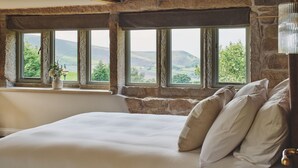 A bedroom with a stone wall, a large bed, and a view of the countryside through multiple windows. - Rye Top Farm - Luxury Cottages (Uppermill)