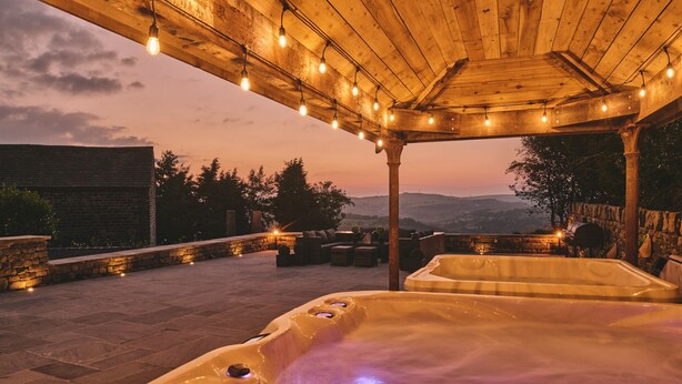 A hot tub area with a wooden pergola overhead, stone walls, and a view of distant hills. - Rye Top Farm - Luxury Cottages (Uppermill)