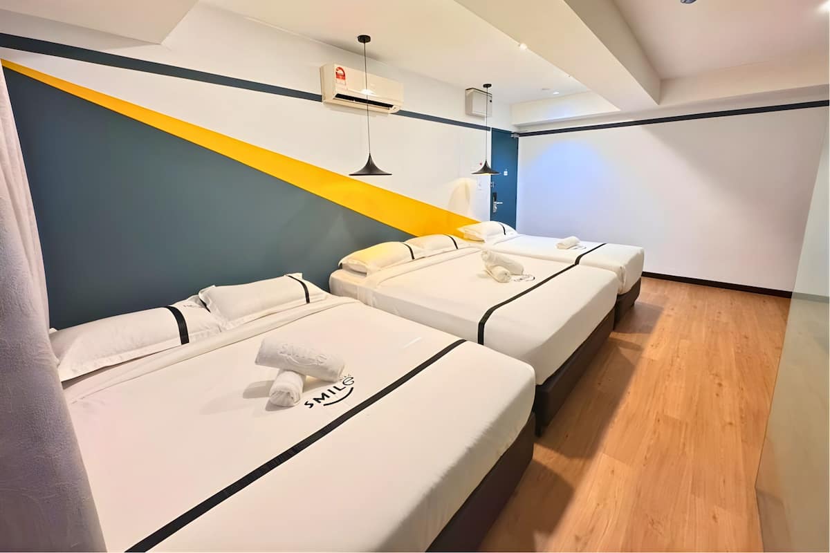 Een hotelkamer met drie bedden, een luchtverkoeler in het plafond, een bureau met stoel en een deur met blauwe omlijsting.
