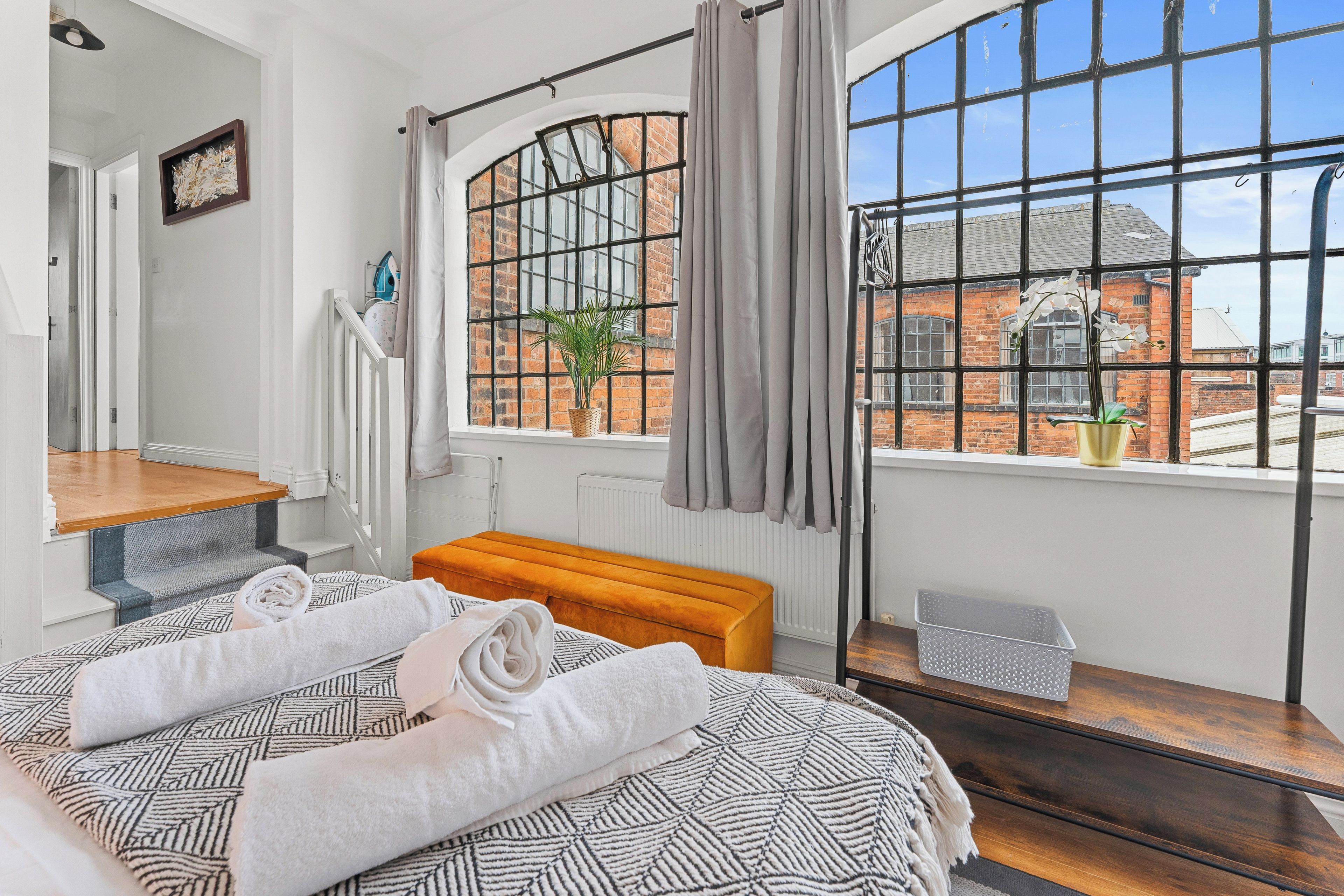 Een slaapkamer met een groot raam, een bed met opgerolde handdoeken, een bankje en een radiator.