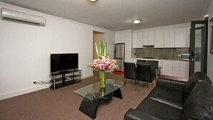 A modern living room with a flat-screen TV, a glass coffee table with a vase of flowers, a black leather sofa, and a dining area with a small kitchen in the background. - One Bedroom Apartment (Newcastle West)