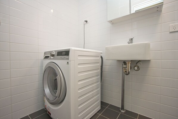 A compact laundry room with a front-loading washing machine, a sink, and tiled walls. - One Bedroom Apartment (Newcastle West)
