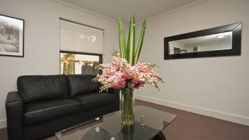 A modern room with a black leather sofa, a glass coffee table with a vase of pink flowers, and a large window with a view of trees.