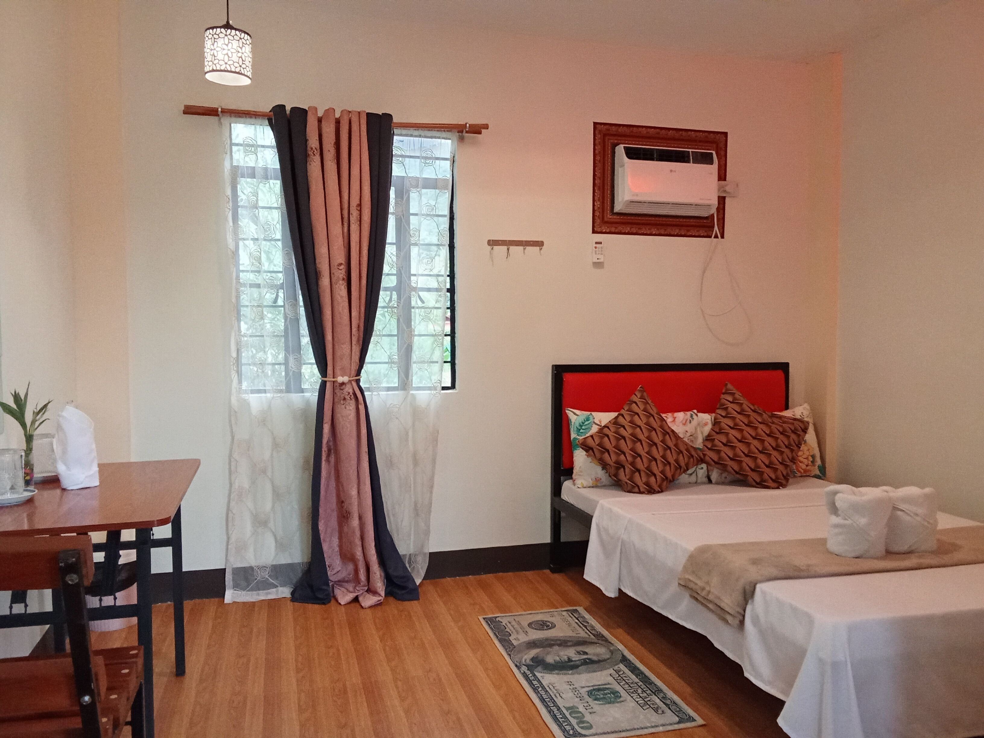 A hotel room with a wooden floor, a bed with a red headboard, a bedside table, a desk with a chair, and a window with curtains.