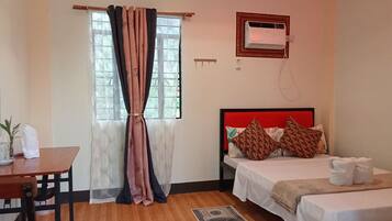 A hotel room with a wooden floor, a bed with a red headboard, a bedside table, a desk with a chair, and a window with curtains.