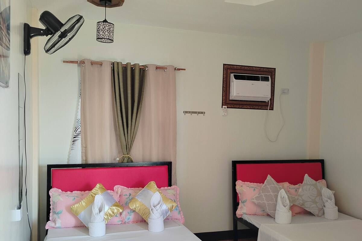 A hotel room with two beds, a ceiling fan, an air conditioning unit, and a window with curtains.