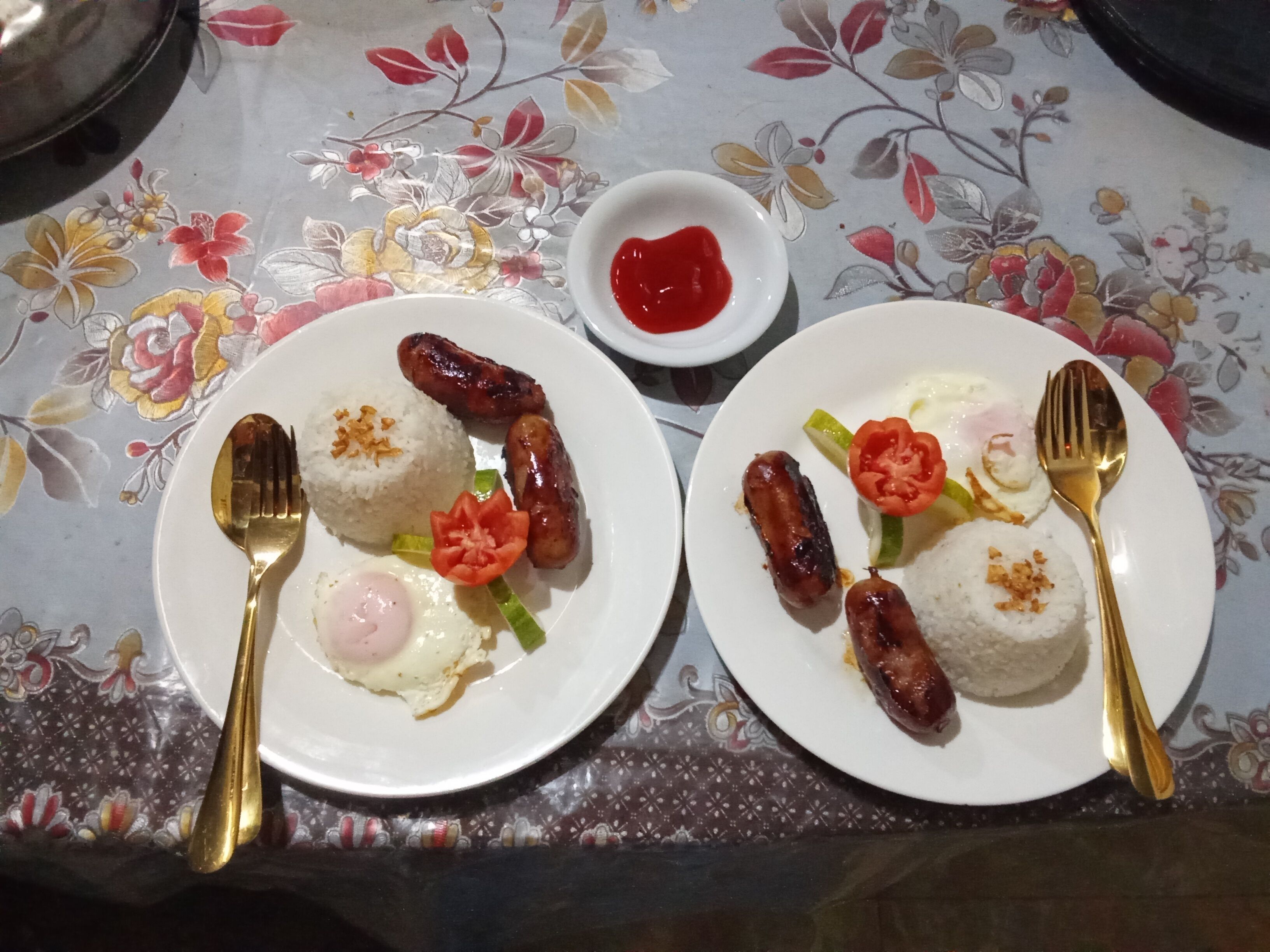 Deux assiettes de nourriture contenant des saucisses, du riz et des œufs, posées sur une table recouverte d’une nappe à motifs floraux.