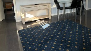 A modern living room with a flat-screen TV mounted on the wall, a light-colored wooden console table, a dark blue patterned rug, and a glass-top dining table with two black chairs. - One Bedroom Disability Access (Cardiff)