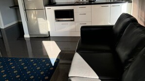 A modern kitchen with white cabinets, a stainless steel refrigerator, a microwave, and a gas stove. There is a black leather sofa and a blue patterned rug in the living area. - One Bedroom Disability Access (Cardiff)