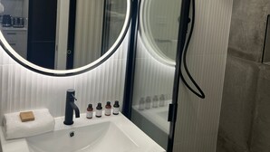 Uma casa de banho moderna com lavatório branco, espelho redondo com iluminação, cabine de duche em vidro e sanita.