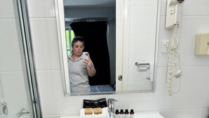 Ein Badezimmer mit weißem Waschbecken, einem Spiegel, einer Dusche und einer Toilette. Auf dem Waschbeckenbrett liegen Kosmetikartikel.