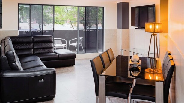 A modern living room with a black leather sofa, a glass dining table with chairs, a TV mounted on the wall, and a large window with a view of the outdoors.