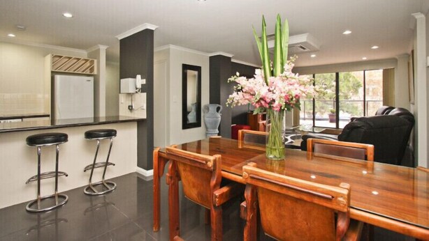 A modern kitchen with a dining area, featuring a wooden table, bar stools, and a vase with flowers. - Two Bedroom Apartment (Newcastle)