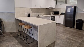 A modern kitchen with white cabinets, a stainless steel refrigerator, a microwave, and a coffee maker. There are two wooden stools at the island. - Two Bedroom Apartment (Newcastle)