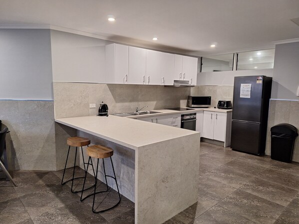 A modern kitchen with white cabinets, a stainless steel refrigerator, a microwave, and a coffee maker. There are two wooden stools at the island.