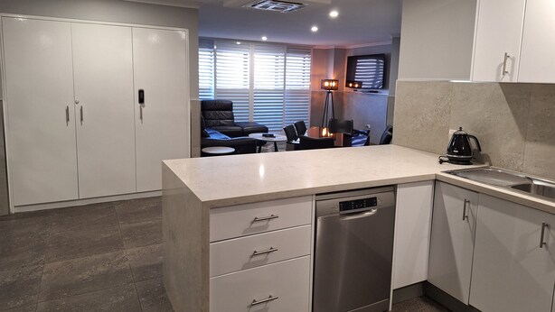 A modern kitchen with a dishwasher, a coffee maker, and a dining area with a table and chairs. - Two Bedroom Apartment (Newcastle)