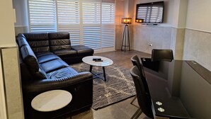 A modern living room with a black leather sofa, a round coffee table, a black dining table with chairs, a standing floor lamp, and a flat-screen TV mounted on the wall. - Two Bedroom Apartment (Newcastle)
