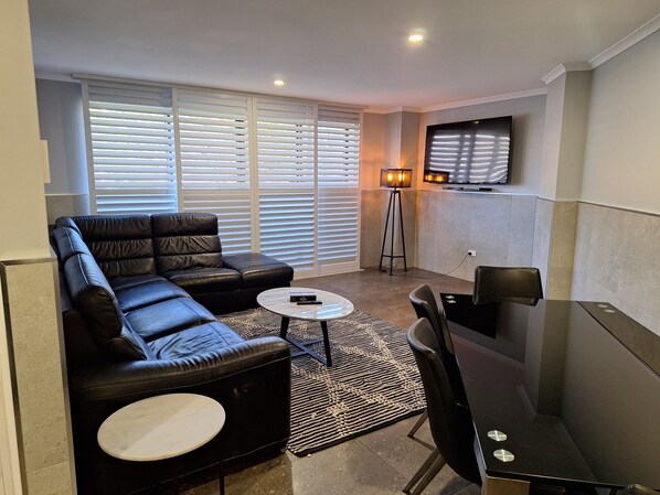 A modern living room with a black leather sofa, a round coffee table, a black dining table with chairs, a standing floor lamp, and a flat-screen TV mounted on the wall.