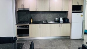 A compact kitchen with light wood cabinets, a black countertop, a stove, a sink, a kettle, and a refrigerator. - Two Bedroom Interconnect (Hamilton)