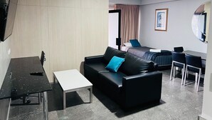 A modern hotel room with a black leather sofa, a small white table, a desk, and a bed with a blue pillow. - Two Bedroom Interconnect (Hamilton)