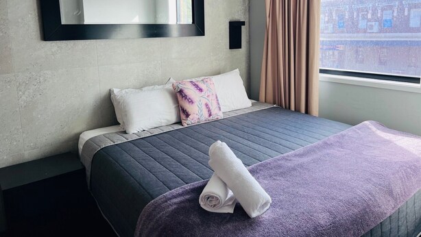 A hotel room with a bed, a large window, a mirror, and a view of buildings outside. - Two Bedroom Interconnect (Hamilton)