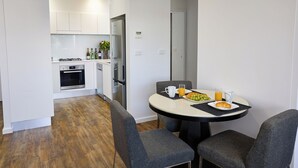 A modern kitchen with white cabinets, a built-in oven, and a refrigerator. There is a dining table with two chairs, a plate of food, a glass of orange juice, and a white mug. - Two Bedroom Apartment (Carrington)