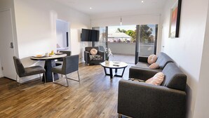 A modern living room with a wooden floor, grey sofas, a dining table with fruit, and a TV mounted on the wall. - Two Bedroom Apartment (Carrington)