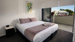 A modern bedroom with a large bed, a nightstand, a painting on the wall, and a view of the outside through a sliding door. - Two Bedroom Apartment (Carrington)