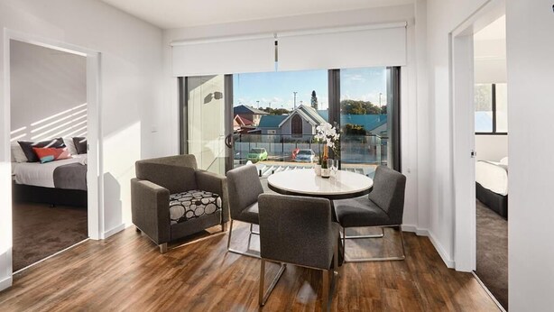 A modern living room with a round table, grey sofas, and a view of a lake and buildings. - Two Bedroom Apartment (Carrington)