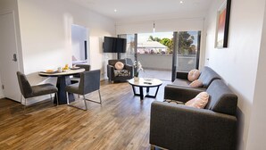A modern living room with a wooden floor, grey sofas, a dining table with fruit, and a TV mounted on the wall. - Two Bedroom Apartment (Carrington)