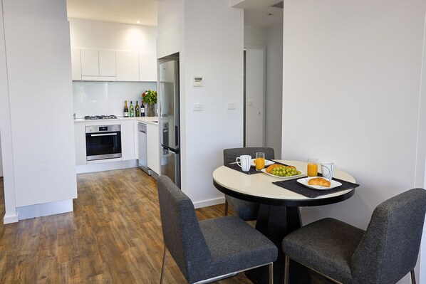 Cocina moderna con armarios blancos, horno empotrado y refrigerador. Hay una zona de comedor con una mesa redonda, dos sillas y una mesa para el desayuno con un croissant, jugo de naranja y una fuente de fruta.
