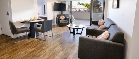 A modern living room with a wooden floor, grey sofas, a dining table with fruit, and a TV mounted on the wall.