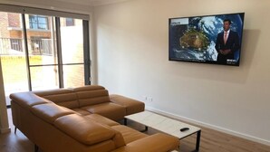 A modern living room with a sectional sofa, a marble coffee table, and a television mounted on the wall displaying a satellite map. - Two Bedroom Apartment (Waratah West)