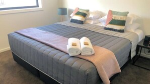 A bed with a grey quilt, a pink blanket, and two rolled towels on the footboard. - Two Bedroom Apartment (Waratah West)