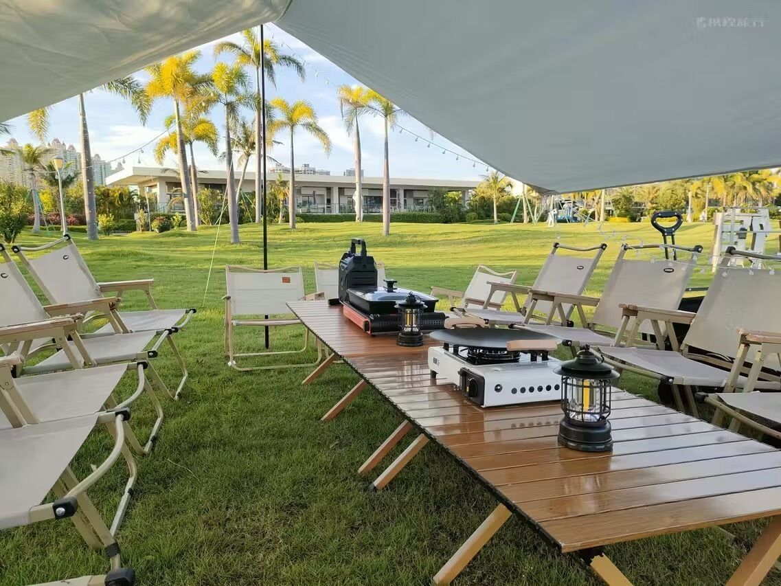 Un espace extérieur aménagé pour les repas, avec un barbecue et des lanternes, entouré de palmiers et avec un b âtiment en arrière-plan.