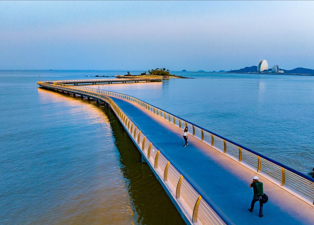 Een lange voetgangersbrug over het water, met iemand aan het wandelen en een ander die foto's maakt.