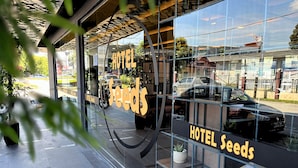 Entrada de un hotel con fachada de vidrio, un letrero que dice "HOTEL Seeds" y el reflejo de un automóvil y edificios.